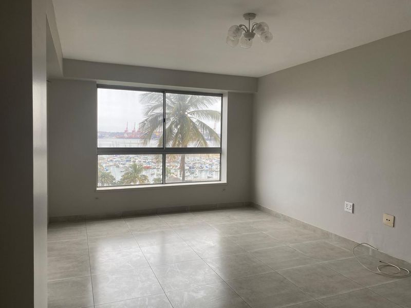 A bright, empty living room featuring large windows with a scenic harbor view and neutral tiled flooring.