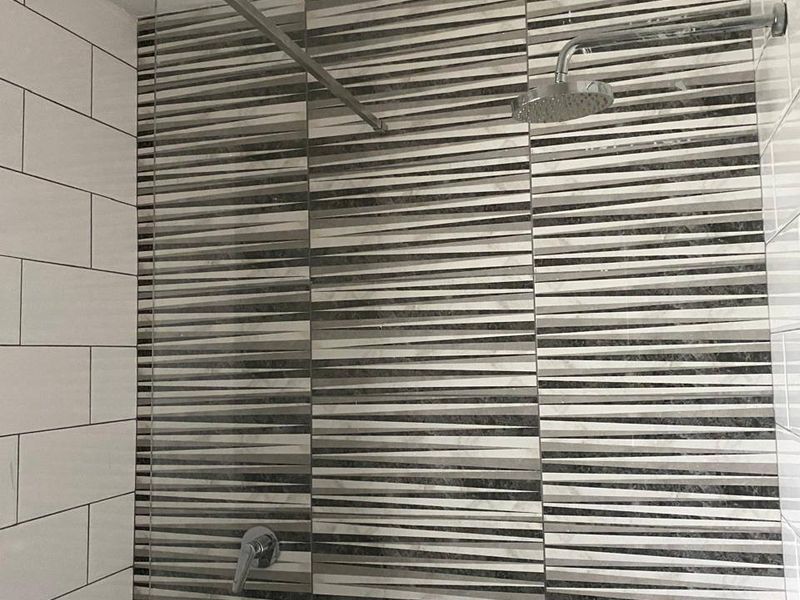 A modern shower enclosure featuring a rainfall shower head, chrome mixer, glass screen, and decorative striped accent wall tiling.