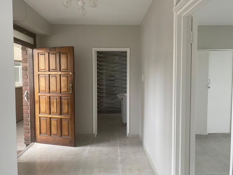 A bright and neutral entrance hall featuring tiled flooring and a solid wood front door, leading into the apartment interior.