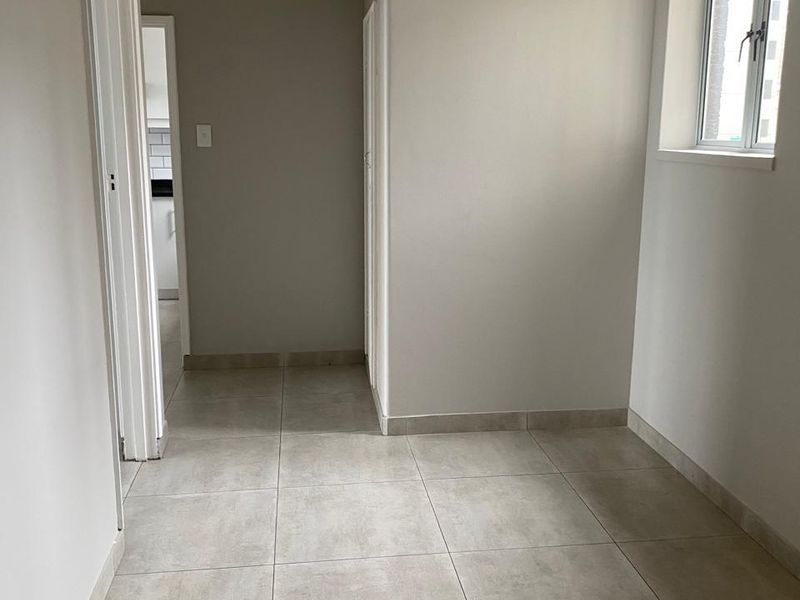 A clean, empty hallway featuring neutral tiled flooring, light-colored walls, and a window providing natural light, leading towards an adjacent room.