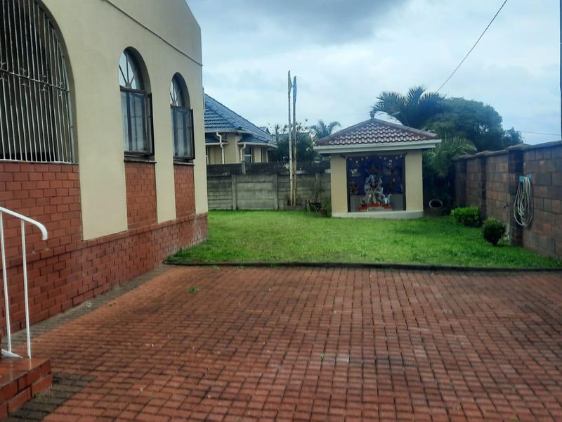 A rear exterior view featuring a paved courtyard, a small garden structure, and a brick boundary wall alongside the main house structure.