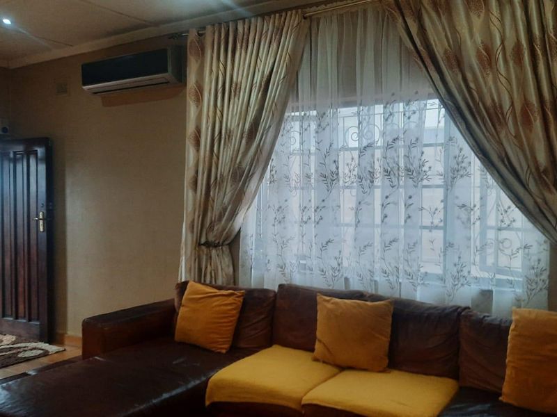 A comfortable living room featuring tiled flooring, a wall-mounted air conditioning unit, and a large window with security bars and curtains.