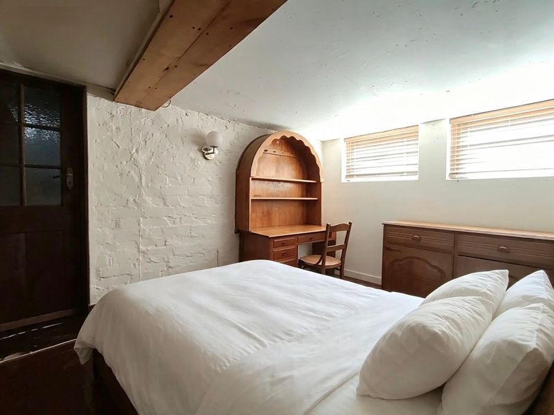 A cozy bedroom featuring an exposed white-painted brick wall, a rustic timber ceiling beam, and natural light through dual windows with venetian blinds.