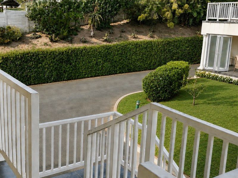 An elevated balcony view overlooking a well-maintained garden, paved driveway, and the exterior facade of the adjacent building section.