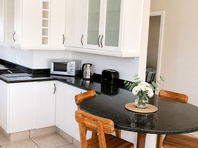 Open-plan kitchen featuring white cabinetry, dark granite countertops, integrated oven, and hob, seamlessly flowing into the dining area with tiled flooring throughout.