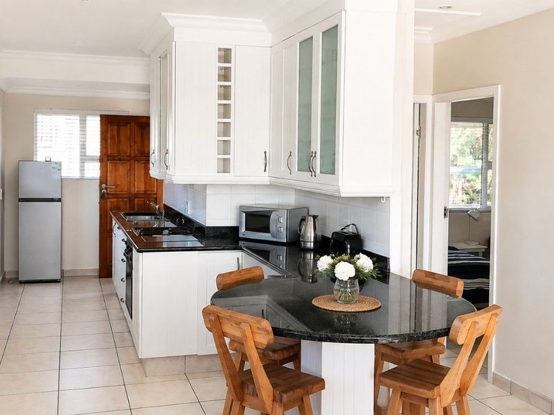 Open-plan kitchen and dining area featuring white cabinetry, granite countertops, tiled flooring, and natural light through venetian blinds.