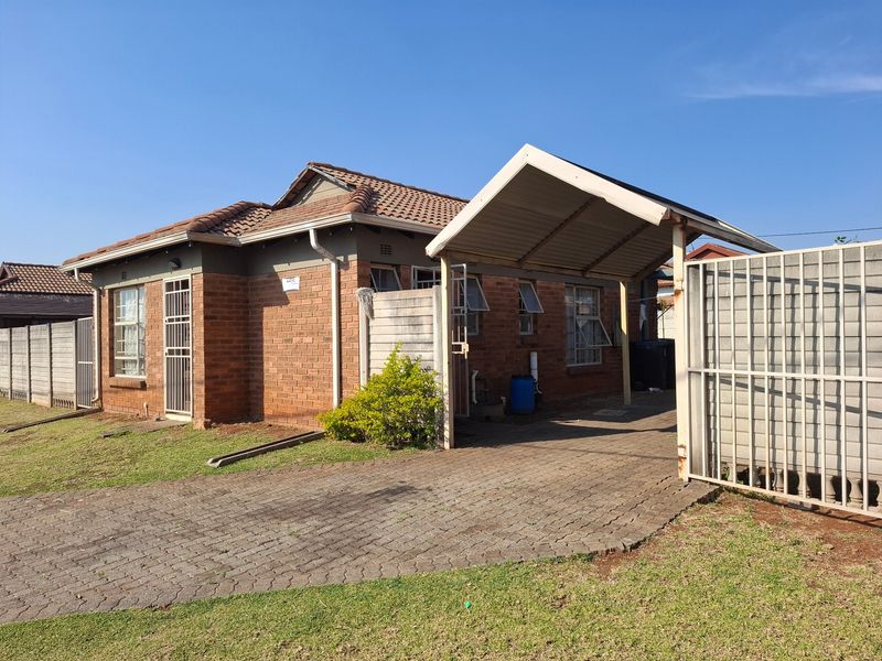 Front view of a single-story brick house featuring a paved driveway, attached carport, and secure perimeter fencing.