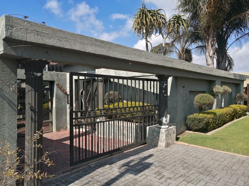 Front entrance featuring a metal security gate, paved driveway, and a concrete boundary wall with decorative columns.