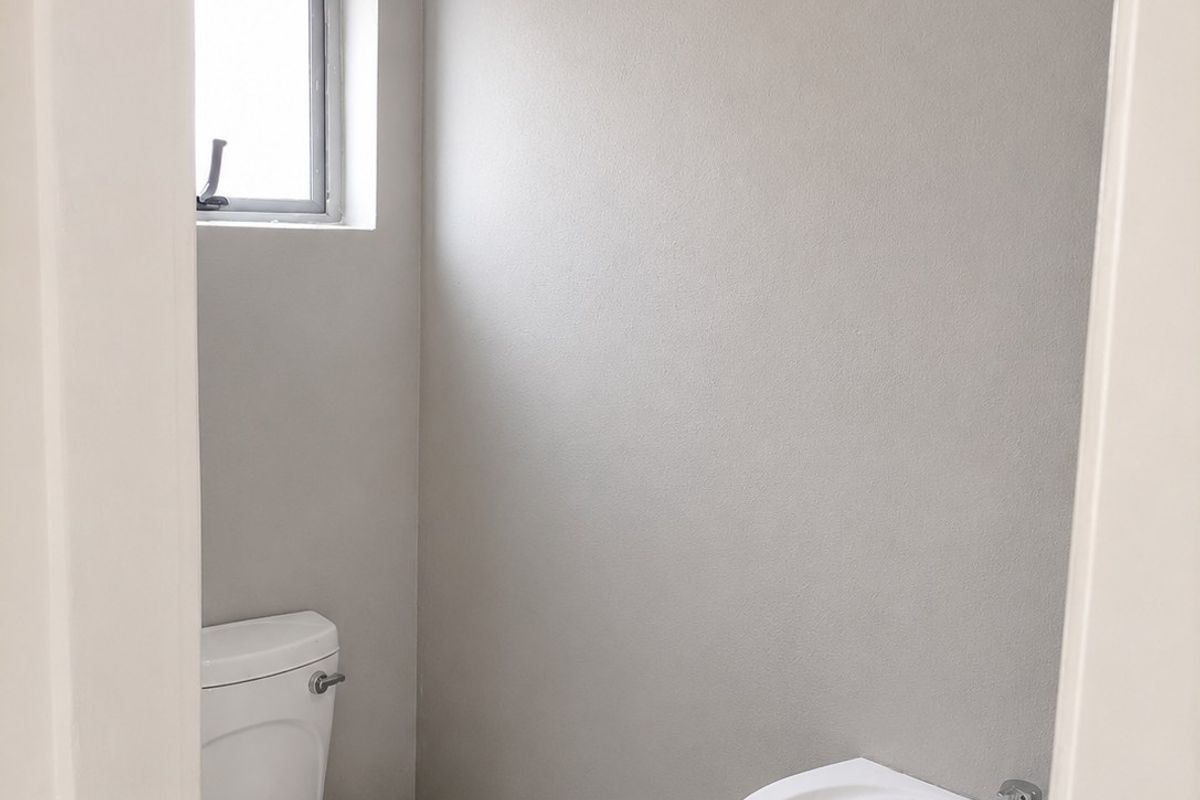 A clean, minimalist guest bathroom featuring a white pedestal basin, toilet, and neutral grey wall finishes with tiled flooring.