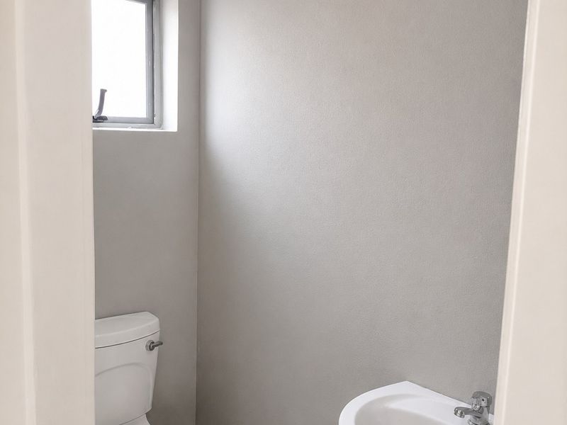 A clean, minimalist guest bathroom featuring a white pedestal basin, toilet, and neutral grey wall finishes with tiled flooring.