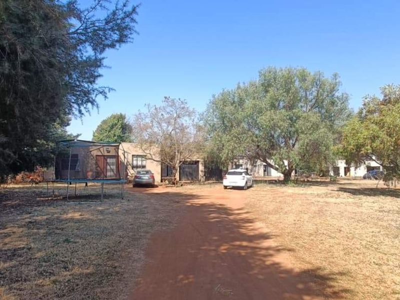 A wide-angle view of the property entrance featuring a dirt driveway leading to the main building, surrounded by mature trees and open land.