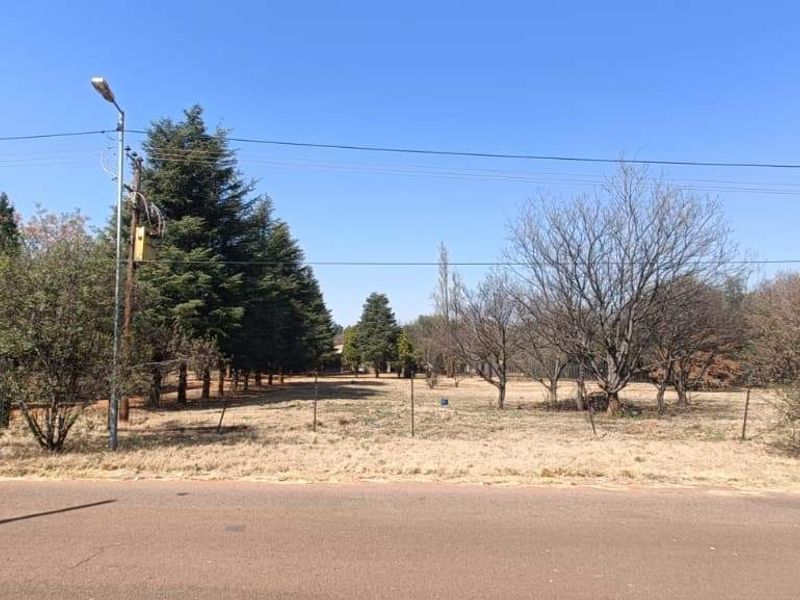 A street-level view of a vacant farm plot featuring established trees, perimeter fencing, and access gate.