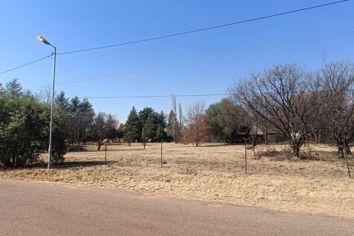 A street-level view of a vacant farm plot featuring wire fencing, mature trees, and existing utility infrastructure along the roadside.