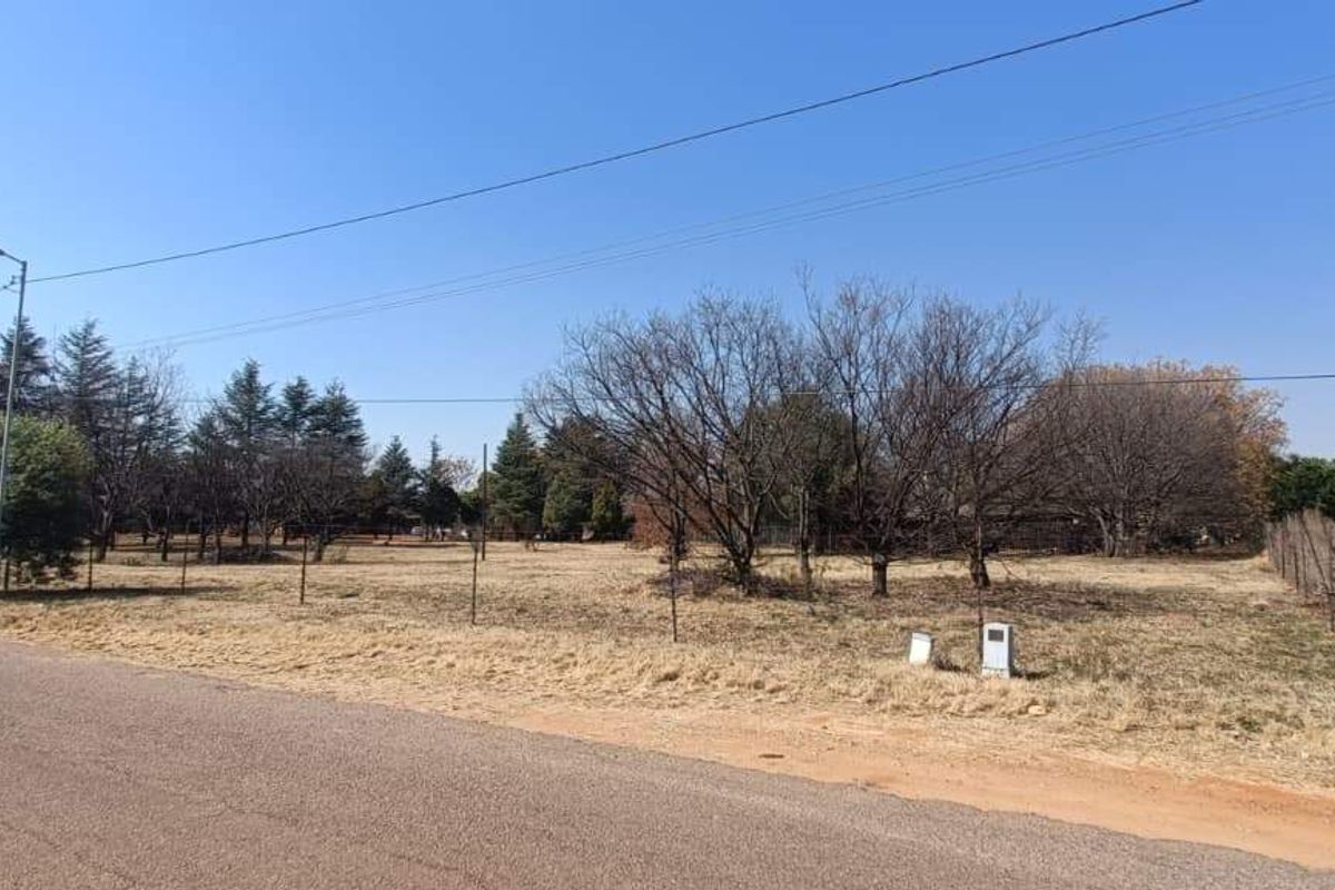 A street-level view of a vacant farm plot featuring established trees, wire fencing, and access to a paved road.