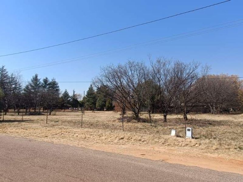 A street-level view of a vacant farm plot featuring established trees, wire fencing, and access to a paved road.