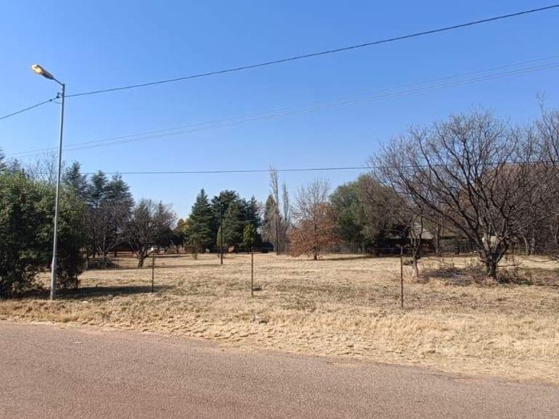A street-level view of a vacant farm plot featuring wire fencing, mature trees, and existing utility infrastructure along the roadside.