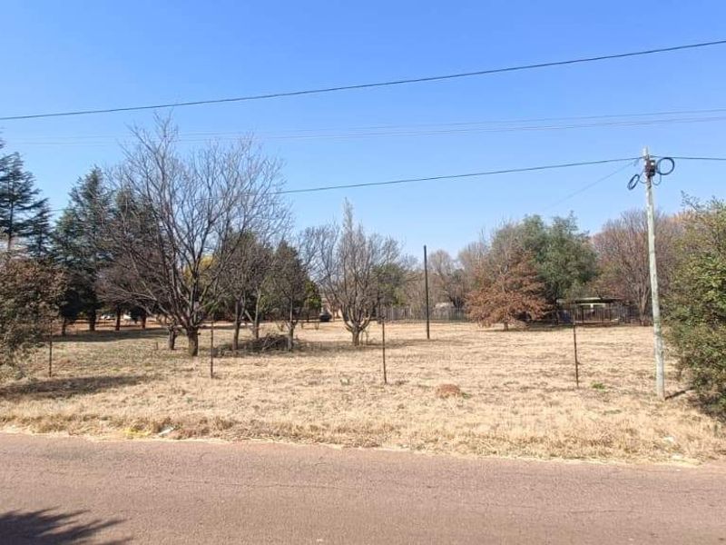 A street-level view of a vacant farm plot featuring established trees, wire fencing, and utility infrastructure.