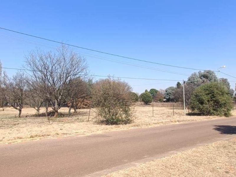 A street-level view of the property frontage, featuring an asphalt road, wire fencing, and open land with established trees.