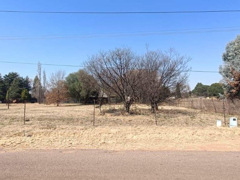 A street-level view of a vacant farm plot featuring dry grass, mature trees, and perimeter wire fencing along the road frontage.