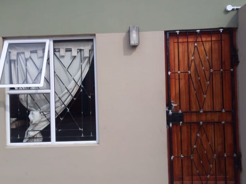 Front exterior view featuring a wooden door with a security gate, an aluminium window with security bars, and an external wall light.