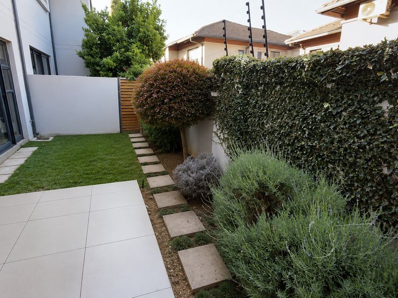 A well-maintained rear garden featuring a tiled patio, manicured lawn, stone pathway, and established greenery against a secure boundary wall.
