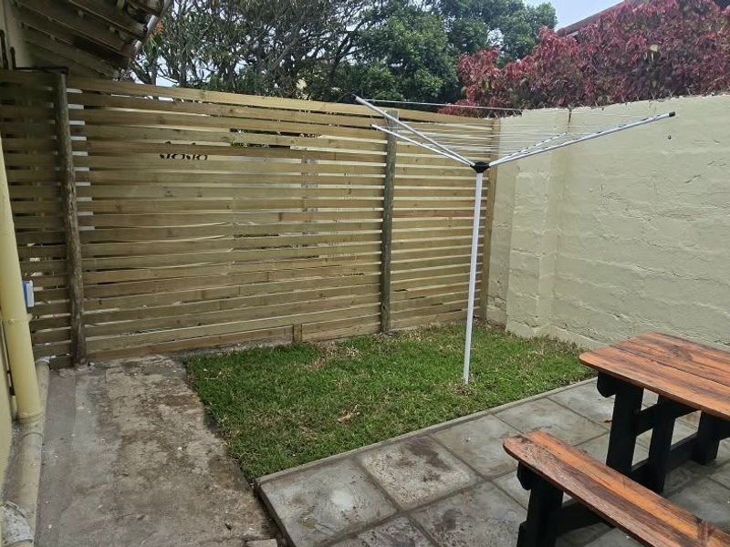 A private rear courtyard featuring a paved patio, small lawn area, and timber privacy screening, enclosed by a masonry boundary wall.
