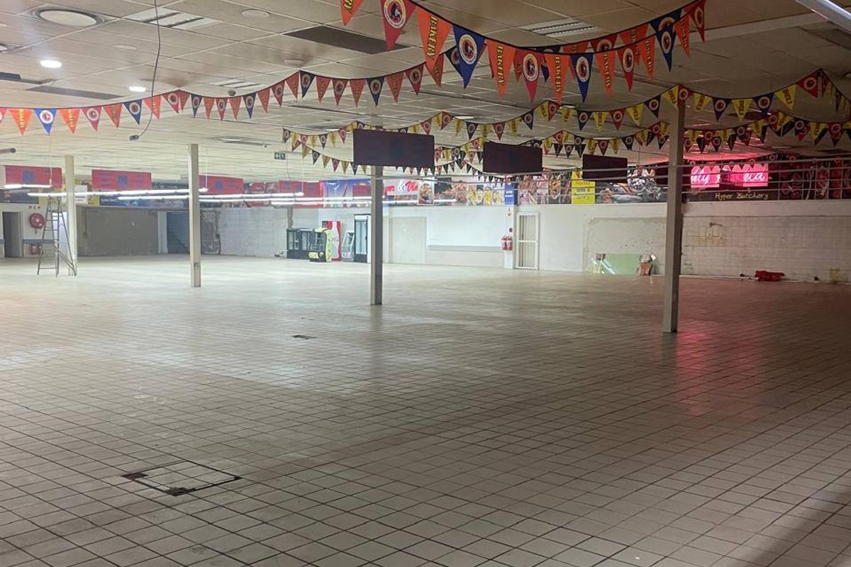 A large, vacant commercial open-plan retail space featuring tiled flooring, a suspended ceiling with recessed lighting, and structural support columns.