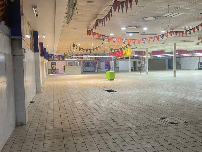 A large, vacant commercial open-plan retail space featuring tiled flooring, a suspended ceiling with recessed lighting, and structural support columns.