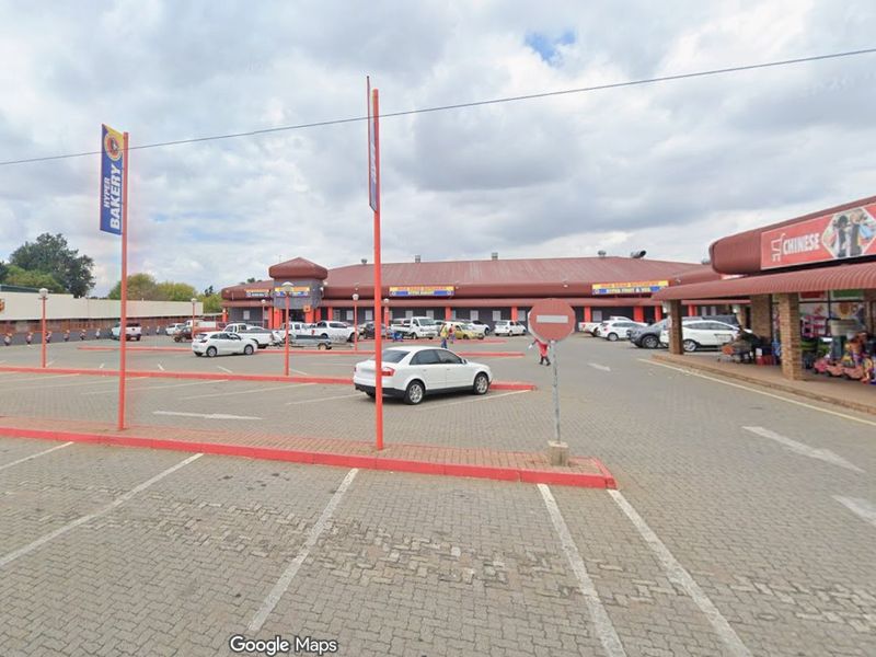 A commercial retail center featuring a large paved parking lot, covered walkways, and multiple storefronts under a continuous corrugated metal roof.