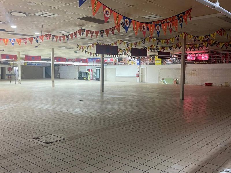 A large, vacant commercial open-plan retail space featuring tiled flooring, a suspended ceiling with recessed lighting, and structural support columns.