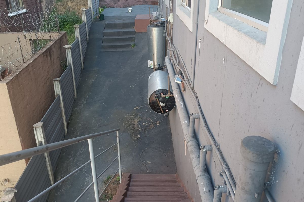 Rear exterior view of the apartment building showing external plumbing, wall-mounted water geysers, concrete stairs, and perimeter fencing.