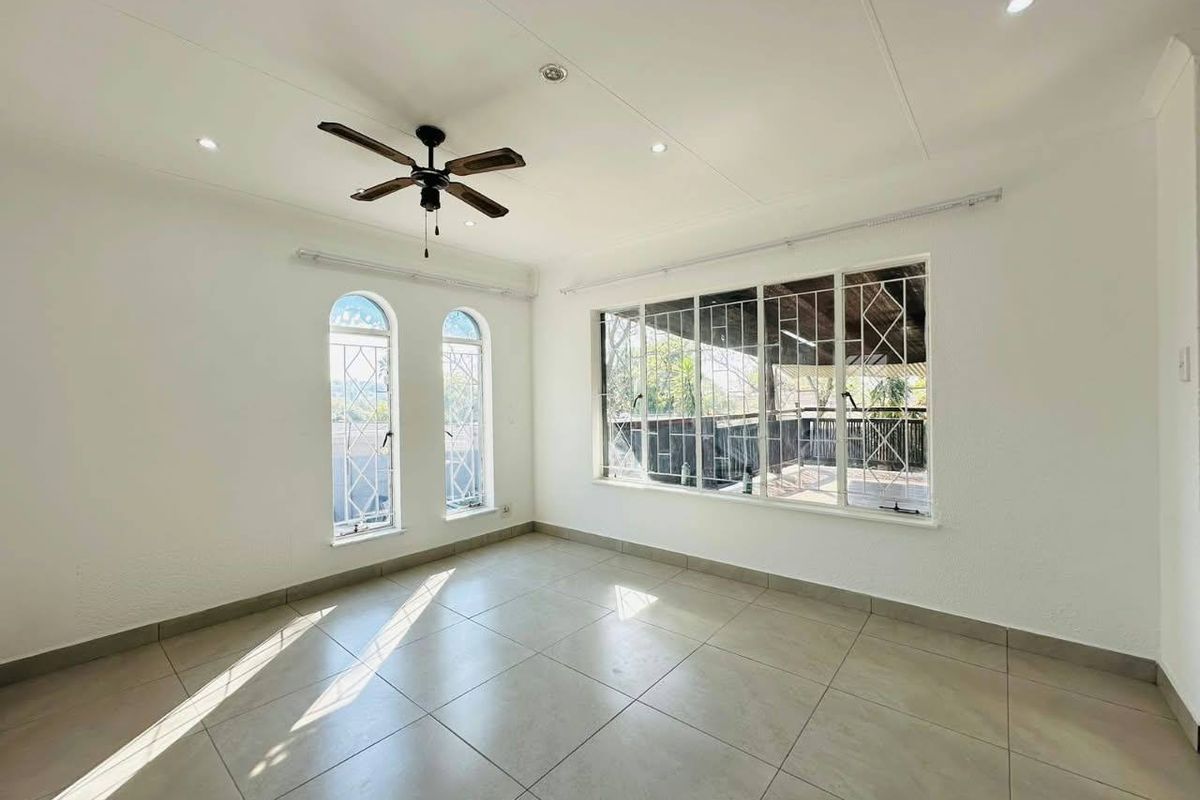 A bright, empty living room featuring tiled flooring, recessed lighting, a ceiling fan, and large windows with security bars overlooking a patio.