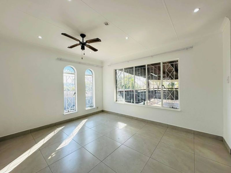 A bright, empty living room featuring tiled flooring, recessed lighting, a ceiling fan, and large windows with security bars overlooking a patio.