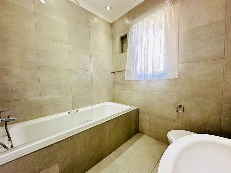 A modern, neutral-toned bathroom featuring a built-in bathtub with a glass screen, tiled walls, and recessed lighting.