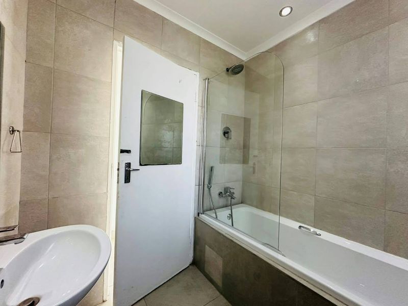 A functional bathroom featuring a bathtub with a glass shower screen, wall-mounted basin, and neutral wall tiling.