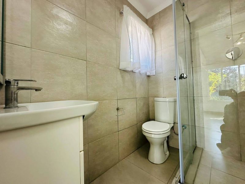 A clean, modern bathroom featuring neutral wall tiles, a vanity basin with chrome mixer, a ceramic toilet, and a glass-enclosed shower.