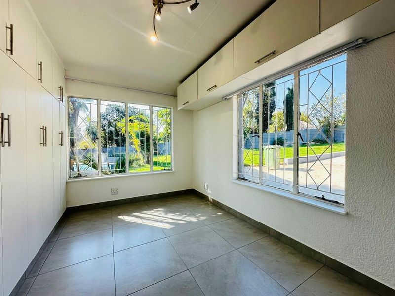 A bright, empty bedroom featuring tiled flooring, extensive built-in cupboard storage, and large windows with security bars overlooking the garden.