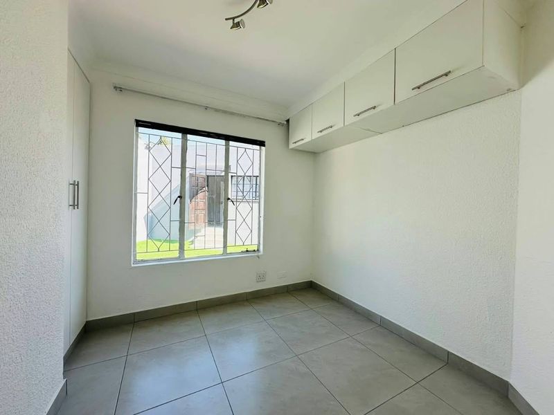 A clean, empty bedroom featuring neutral tiled flooring, built-in cupboards, overhead storage cabinets, and a window fitted with security bars.
