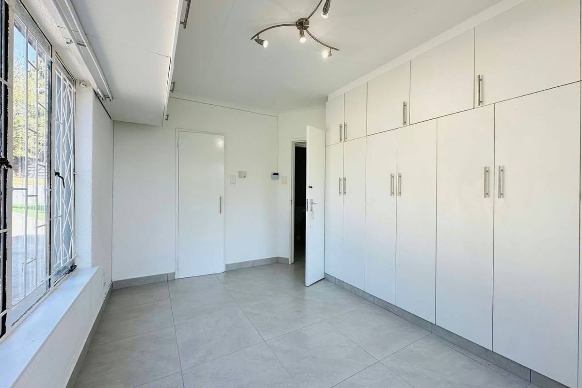 A clean, empty bedroom featuring neutral tiled flooring, extensive built-in storage cupboards, and security bars on the window.