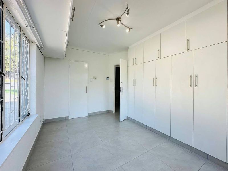 A clean, empty bedroom featuring neutral tiled flooring, extensive built-in storage cupboards, and security bars on the window.
