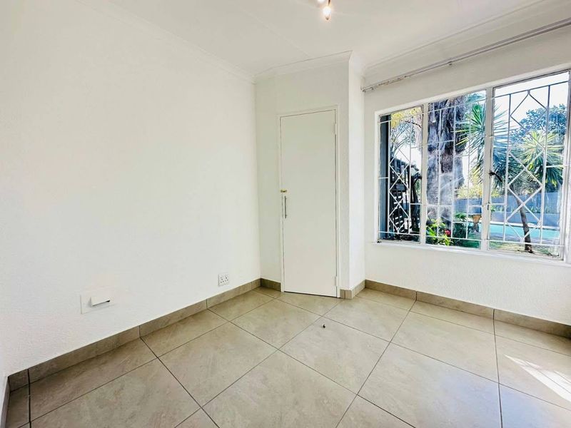 A bright, empty bedroom featuring neutral tiled flooring, a built-in cupboard, and large windows fitted with security burglar bars overlooking the garden.