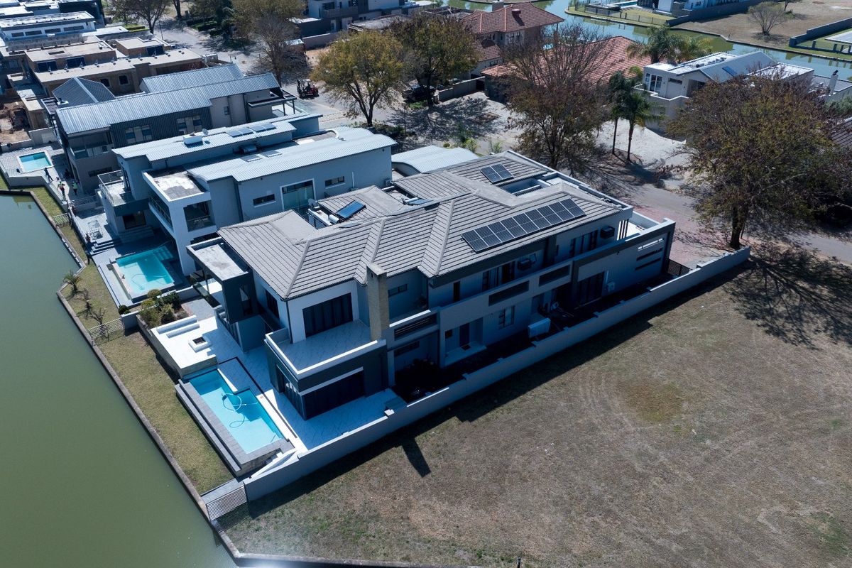 Aerial view of a modern waterfront property featuring a tiled roof with solar panels, a private swimming pool, and a paved patio area.