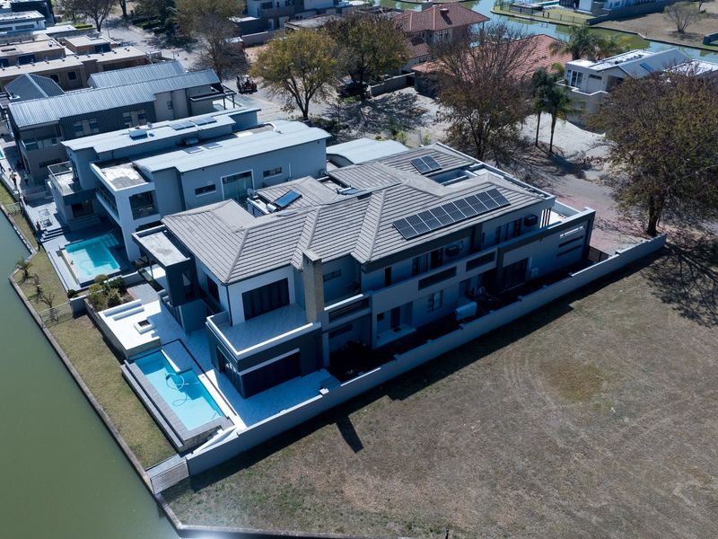 Aerial view of a modern waterfront property featuring a tiled roof with solar panels, a private swimming pool, and a paved patio area.