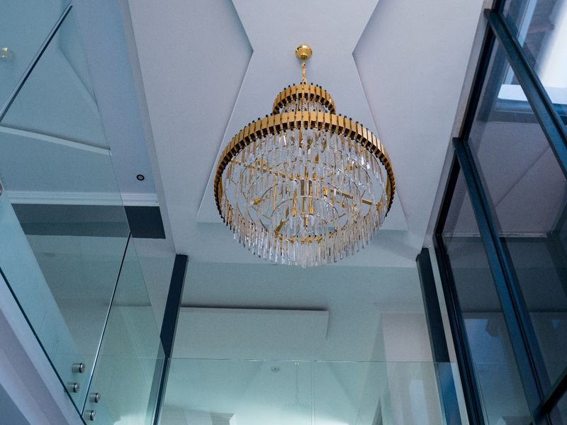A modern, high-ceilinged hallway featuring a striking crystal chandelier, glass balustrades, and clean architectural lines.