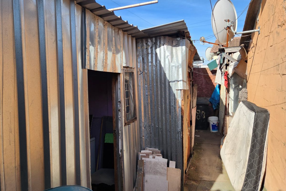A narrow exterior rear passage featuring corrugated metal structures and a concrete pathway, showing significant signs of wear and structural deterioration.