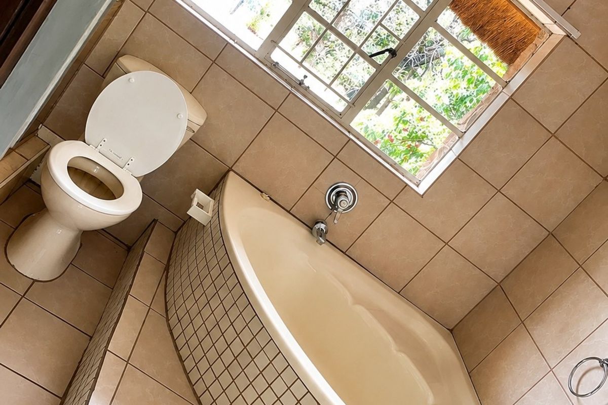 A functional bathroom featuring a built-in corner bathtub, ceramic tiling throughout, and a window with security bars, offering a classic layout ready for modernization.