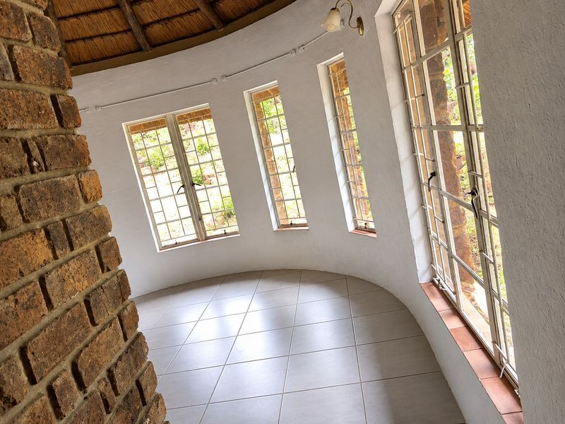 A bright, circular living area featuring exposed brickwork, a traditional thatched roof, tiled floors, and large steel-framed windows overlooking the garden.