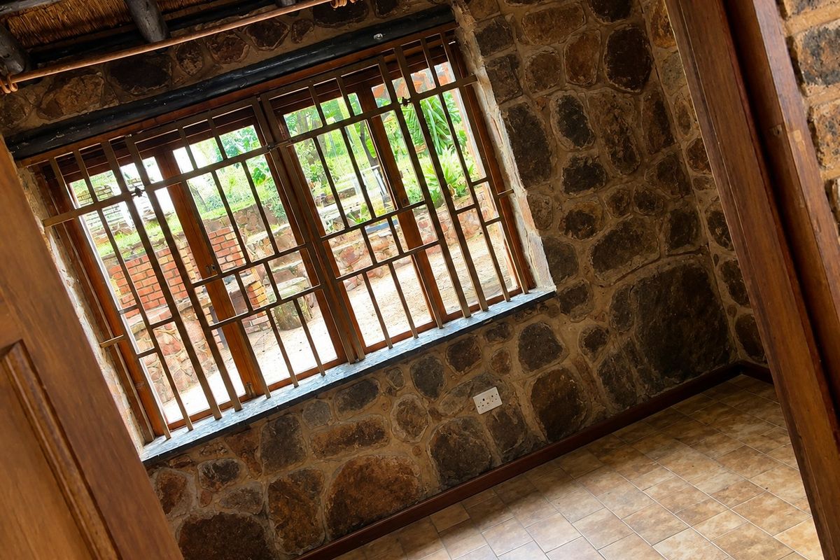 An empty bedroom featuring rustic stone walls, a thatched ceiling, and timber-framed windows with security bars, finished with ceramic floor tiles.