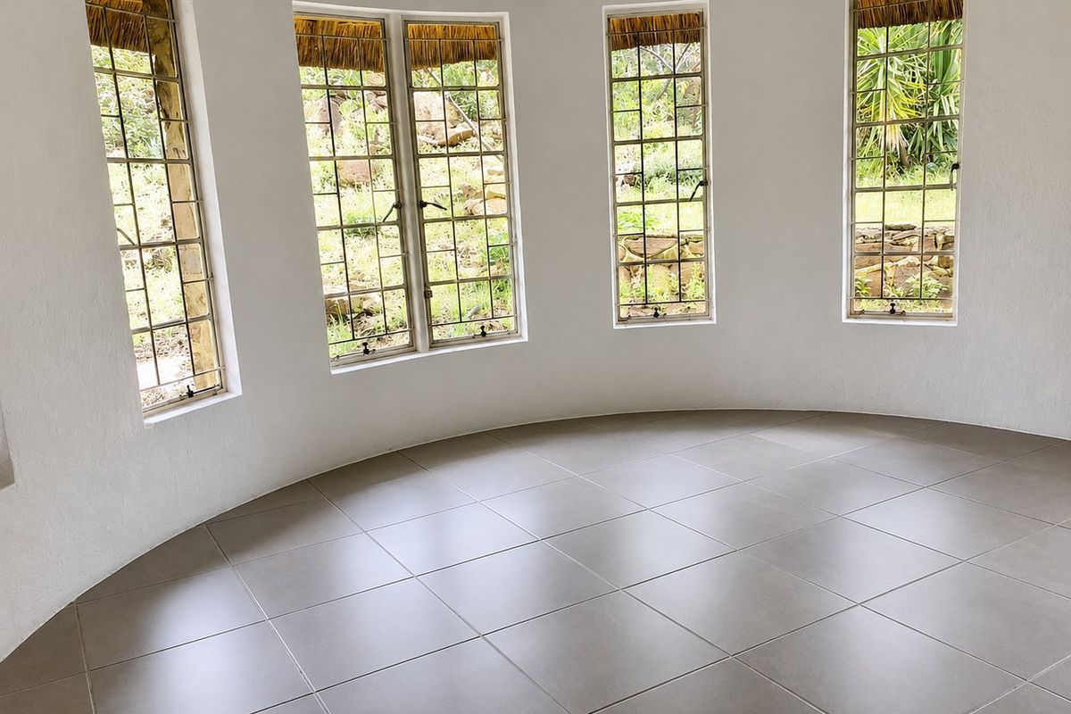 A bright, empty circular living space featuring large steel-framed windows and modern neutral floor tiles, offering a unique architectural layout.