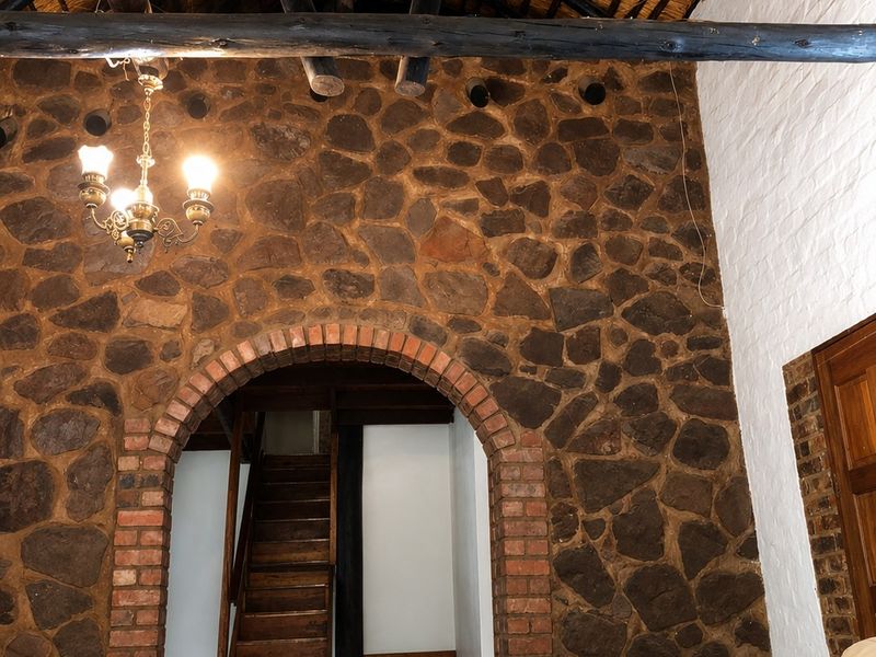 Spacious living area featuring a striking stone feature wall, brick archway, exposed timber rafters, and a high thatched ceiling with a staircase.